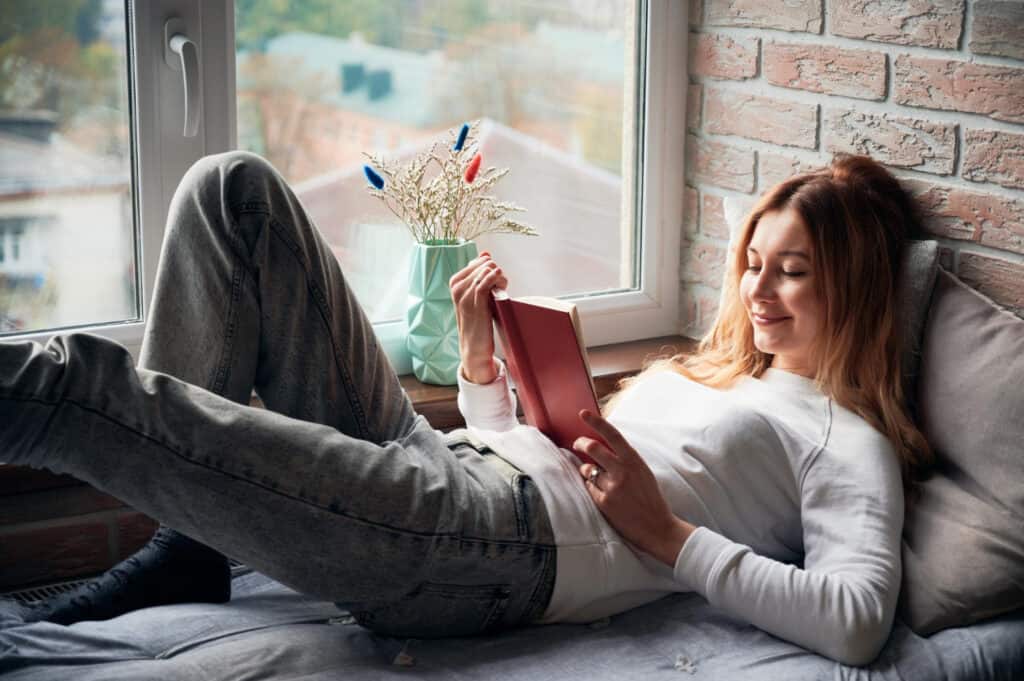 woman is lying in windowsill bed and reading a book